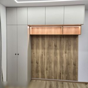 Шкаф Г-образной формы до потолка над кроватью в спальне с подсветкой открытых полок и щитом ЛДСП у изголовья для защиты стены